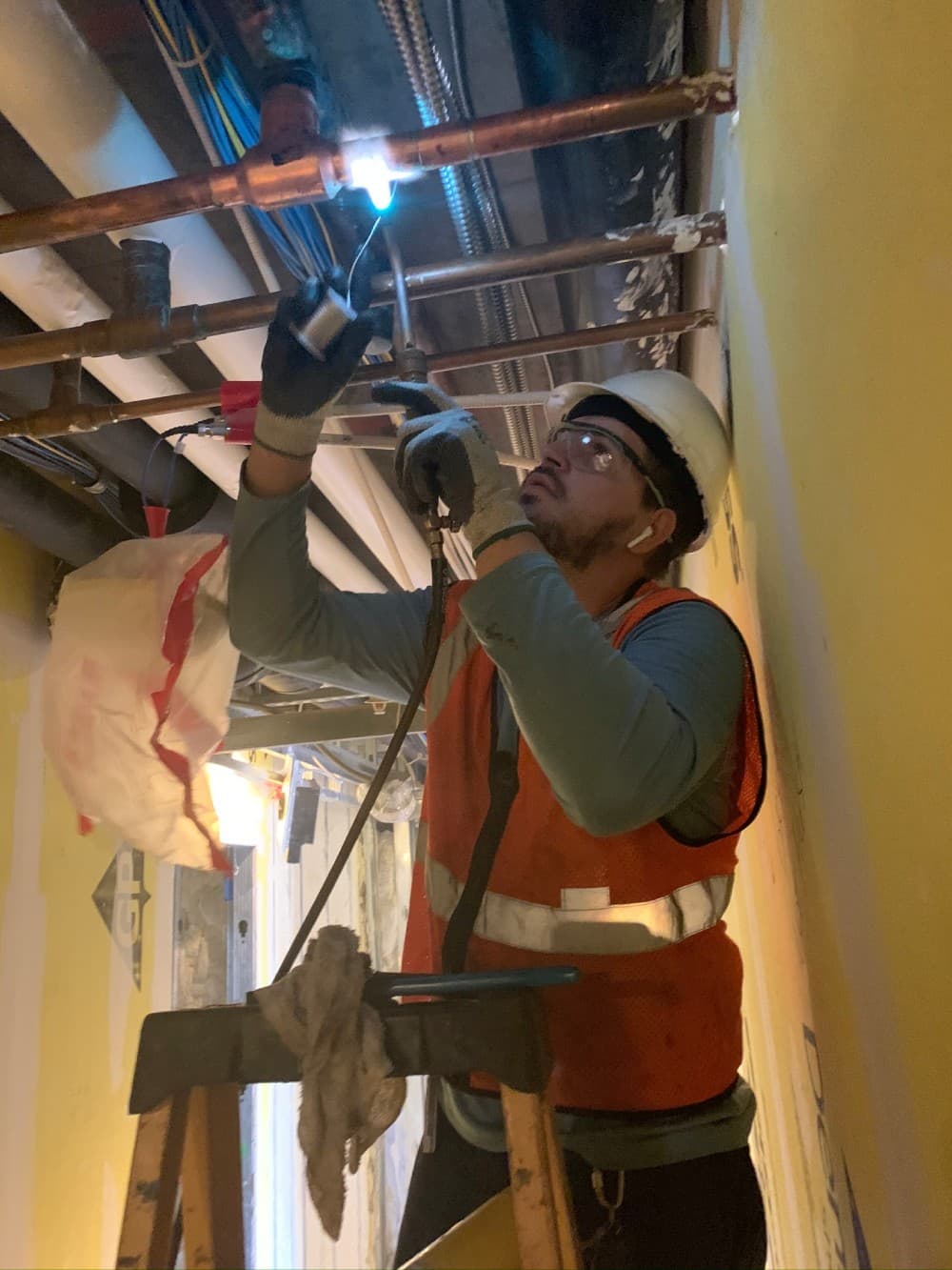 Omnia Plumbing technician in hard hat and high-visibility vest performing overhead plumbing work on a commercial job site.